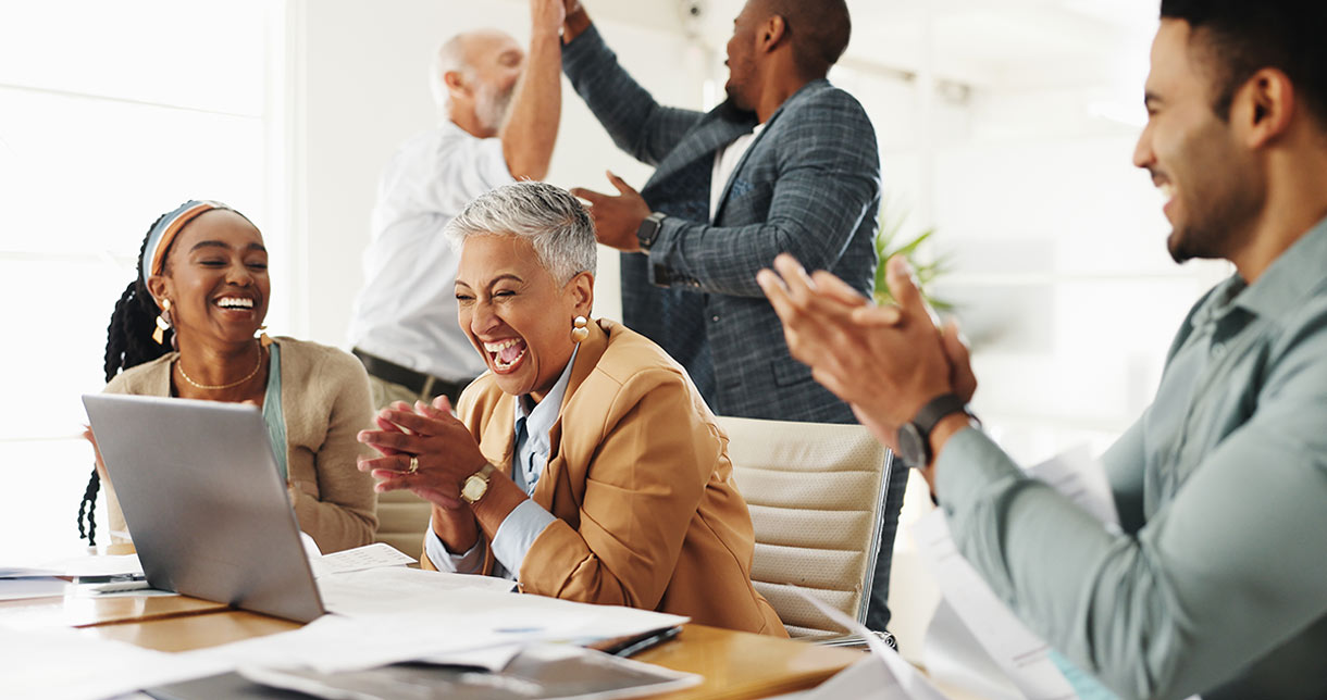 A group of diverse colleagues celebrate at a desk, smiling, laughing, clapping, and high-fiving in a brightly lit office setting, with papers and a laptop on the table.