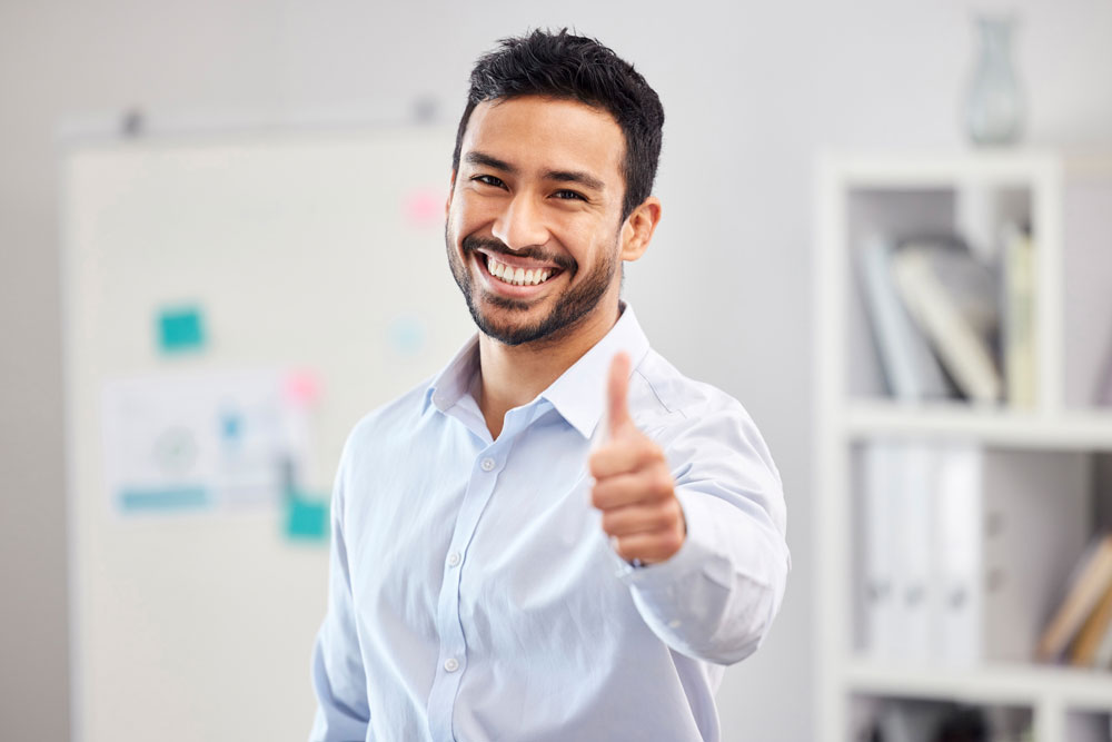 A smiling man with dark hair and a beard, wearing a light blue shirt, stands indoors giving a thumbs up. The background shows a whiteboard with notes and shelves with books and folders.