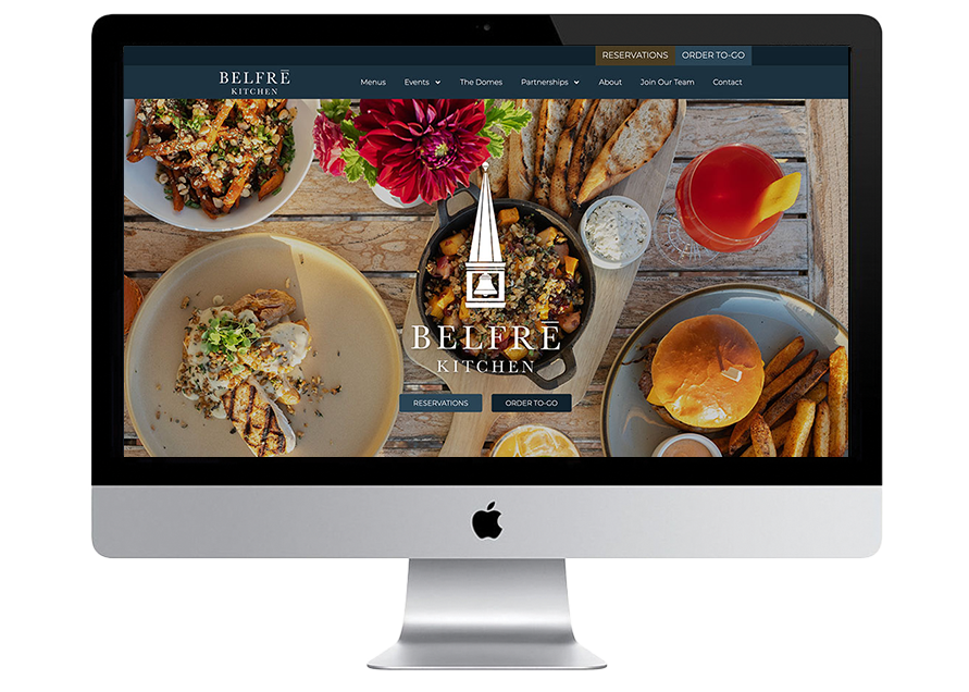 A computer screen displays the homepage of Belfrē Kitchen’s website, featuring plates of food, drinks, flowers, and navigation options like Menu, Events, and Reservations at the top.